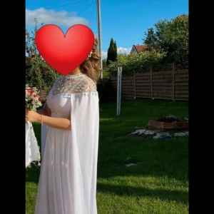 Vit långklänning med cape och pärldetaljer - Elegant vit långklänning med cape och vackra pärldetaljer över axlar och byst. Klänningen har en flytande, luftig passform och är ärmlös med hög halsringning. Perfekt för speciella tillfällen. Använd bara en gång 