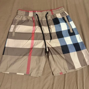 Rutigta badshorts med snörning - Snygga badshorts i klassiskt rutig design med beige, blå, svart och röda detaljer. Shortsen har elastisk midja med svart snörning och sidofickor. Perfekta för stranden eller poolen.