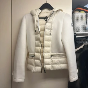 Vit pufferjacka med huva från JOTT - Säljer en snygg vit pufferjacka från JOTT  med huva och dragkedja. Jackan har quiltade partier framtill och baktill, samt två fickor med dragkedja. Ärmarna är släta och har dragkedjedetaljer vid handlederna. Perfekt för kyliga dagar.