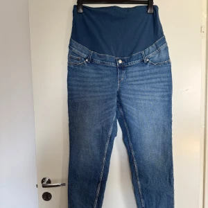 Gravid jeans från H&M, nyskick - Blå mamma jeans från H&M med mjuk, stretchig mudd över magen. Klassisk femficksmodell med raka ben och normal passform. Perfekta för dig som vill ha bekväma jeans under graviditeten. Väldigt sparsamt använda, nyskick. Tvättade. Djurfritt och rökfritt hem.