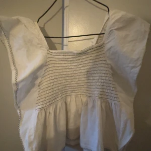 Vit blus med smock från Zara - Säljer en vit blus från Zara med smockad överdel och volang nedtill. Blusen har korta, vida ärmar och dekorativ söm längs kanterna. Perfekt för en somrig och luftig stil.