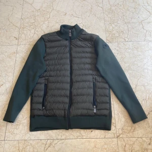 Mörkgrön hybridjacka från Moncler - Snygg mörkgrön jacka från Moncler med quiltad front och släta ärmar. Jackan har dragkedja framtill, två fickor med dragkedja och hög krage. Perfekt för lager-på-lager och stilren look.