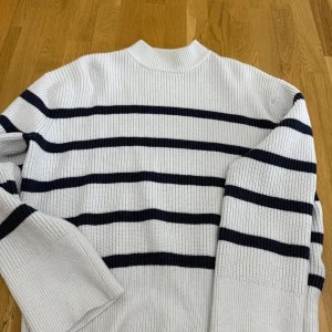 Vit och marinblå randig stickad tröja - Säljer en vit stickad tröja med marinblå horisontella ränder. Tröjan har lång ärm och ribbad struktur, perfekt för kyligare dagar. Klassisk och stilren design som passar till mycket.