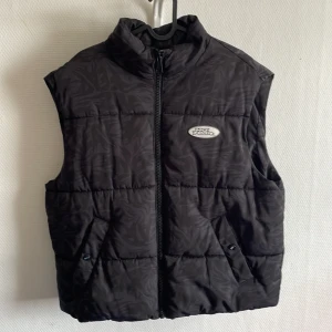 Svart dunväst från No Fear x H&M - Svart dunväst med diskret mönster och hög krage från No Fear x H&M. Västen har dragkedja framtill, två snedställda fickor med knapp och en broderad logotyp på bröstet. Perfekt för lager-på-lager-stil.