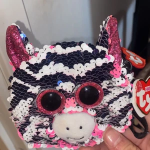 Zebramönstrad plånbok med paljetter från Ty - Supergullig plånbok från Ty i form av ett zebrahoved med svart, vit och rosa paljettmönster. Stora glittriga rosa ögon och öron, dragkedja upptill. Perfekt accessoar för att förvara småsaker eller mynt.