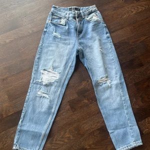 Ljusblå mom jeans från Amisu - Säljer ett par ljusblå mom jeans från Amisu med slitna detaljer och hål på benen. Jeansen har hög midja, raka ben och klassisk femficksdesign. Perfekta för en avslappnad och trendig look.