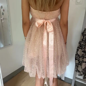 Ljusrosa kortklänning med rosett från Ida Sjöstedt - Supersöt ljusrosa kortklänning från Ida Sjöstedt med tunna axelband och stor rosett i midjan. Klänningen har ett transparent tylltyg med små prickar och ett fint fall. Perfekt för speciella tillfällen!