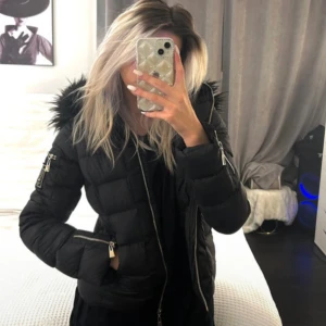 Svart pufferjacka med fuskpäls - Säljer en svart pufferjacka med huva och fuskpälsdetalj. Jackan har dragkedja framtill och på ärmarna, samt quiltad design. Perfekt för kyliga dagar och riktigt snygg till vardags.