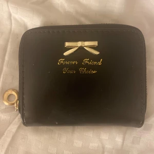 Svart plånbok  - Säljer en svart plånbok med guldfärgad rosett och texten 'Forever Friend Your Choice' på framsidan. Plånboken har dragkedja runtom och flera fack inuti, inklusive ett myntfack med röd dragkedja och prickigt tyg. Vet tyvärr inte vart den är ifrån