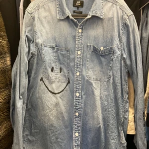 Jeansskjorta med smiley från Lee - Säljer en ljusblå jeansskjorta från Lee med två bröstfickor och en stor svart smiley på framsidan. Skjortan har klassisk krage, knappar framtill och lång ärm. Perfekt för dig som vill sticka ut lite extra! Behöver strykas men annars fin