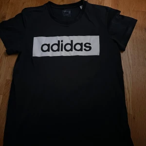 Svart t-shirt från Adidas med logga - Svart t-shirt från Adidas med stor vit logga framtill och liten logga på ryggen. Klassisk passform och rund halsringning. Perfekt för en sportig och avslappnad stil.