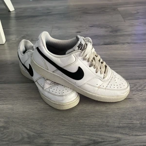 Vita Nike sneakers med svart swoosh - Klassiska vita Nike sneakers med svart swoosh på sidan. Skorna har snörning, rund tå och platt sula. Perfekta för vardagsstilen och passar till det mesta. Diskret Nike-logga på plösen och hälen.