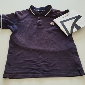 Mörklila pikétröja från Moncler - Säljer en mörklila pikétröja från Moncler med klassisk krage och knappar framtill. Tröjan har vita ränder på krage och ärmslut samt Moncler-logga på bröstet. Perfekt för en stilren och sportig look. Den är köpt på NK för cirka 1800. Kan gå lägre pris via snabb affär. Den passar en person som är runt 152-168. Perfekt för im du vill se grisch ut på sommaren.