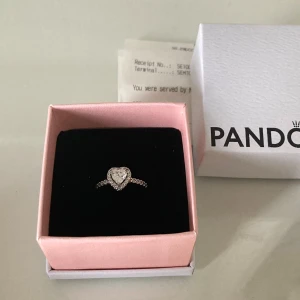 Silverring med hjärta från Pandora - Säljer en elegant silverring från Pandora med ett gnistrande hjärta i mitten, omgiven av små stenar. Ringen levereras i originalask och är perfekt för att ge en romantisk touch till din stil. Storlek 7