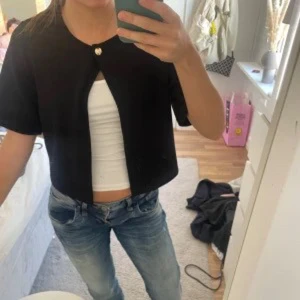 Svart blus från Gina Tricot - Säljer en svart croppad blus från Gina Tricot med rund halsringning och en dekorativ knapp framtill. Jackan har korta ärmar och en stilren, minimalistisk design som passar perfekt till jeans eller kjol.