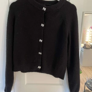 Svart kofta med rosettknappar från Vero Moda - Säljer en svart stickad kofta från Vero Moda i storlek M (Mer likt storlek S). Framsidan pryds av dekorativa silvriga rosettknappar som ger en söt detalj till plagget. Endast använd några gånger💗 Medföljer även en extra rosett💗 