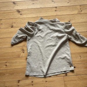 Beige topp med puffärmar från Pernilla Wahlgren - Säljer en beige topp från Pernilla Wahlgren med korta puffärmar och lätt struktur i tyget. Toppen har en liten ståkrage och knäppning bak i nacken. Perfekt för dig som gillar stilrena och trendiga plagg.