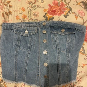 Blå jeantopp med knappar - Säljer en blå jeanstopp med fransig nederkant och knappar framtill. Toppen har två fickor med lock och knappdetaljer samt smock baktill för extra stretch.