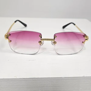 Rosa tonade solglasögon med gulddetaljer - Stilrena solglasögon med fyrkantiga, rosa till transparent tonade glas och guldfärgade skalmar. Glasögonen har minimalistisk design utan båge runt glasen och svarta ändar på skalmarna. Perfekt accessoar för en trendig look.