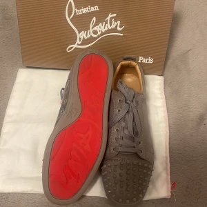 Grå Christian Louboutin sneakers med nitar - Säljer ett par grå sneakers från Christian Louboutin med ikonisk röd sula och coola nitar på tån. Skorna har snörning och är tillverkade i mocka med beige innersida. Perfekt för dig som vill sticka ut med lyxig streetstyle.