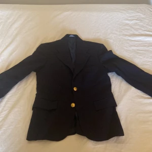 Mörkblå kavaj Polo Ralph Lauren - Klassisk mörkblå kavaj från Polo Ralph Lauren med snygga guldknappar med emblem. Kavajen har två fickor med lock och en stilren passform. Perfekt för dig som vill ha en tidlös och elegant look. Storlek: 10 år, skick: nästan aldrig använd!