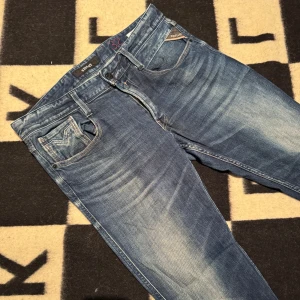 Replay Anbass blå jeans, stl 32/32 - Säljer ett par Replay Anbass jeans i klassisk blå tvätt med snygga slitningar. Jeansen har fem fickor, raka ben och normal passform. Materialet är bomull med lite stretch för extra komfort. Perfekta för dig som gillar en avslappnad men stilren look.