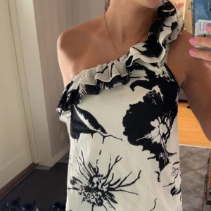 Svartvit blommig oneshoulder topp - Snygg topp med stora svarta blommor på vit botten och volangdetalj längs den ena axeln. Oneshoulder-modell som ger en cool och trendig look. Bara att kontakta mig vid frågor💓💓