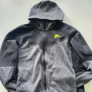 Grå Nike hoodie med dragkedja - Nike hoodie i två nyanser av grått med hel dragkedja och huva. Snygg detalj med neongul Nike-logga på bröstet och en extra ficka med dragkedja på ärmen. Perfekt för chill eller träning, riktigt skön och stilren.