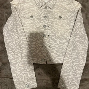 Jättecool jeansjacka - Snygg beige jeansjacka med ljusgrått zebramönster och klassisk krage. Jackan har knäppning framtill, två bröstfickor med knappar och långa ärmar. Perfekt för dig som vill sticka ut med en unik look.