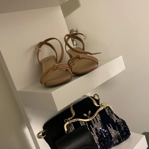 Beiga klackskor  - Snygga beiga klackskor med remmar som korsar framtill. Skorna har öppen tå och justerbart spänne vid ankeln. Tillverkade i glansigt syntetmaterial som ger en stilren look. Perfekta för varmare dagar och när du vill lyfta din outfit.