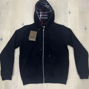 Burberry hoodie svart med dragkedja - Svart hoodie från Burberry med klassiskt rutigt foder i huvan. Tröjan har dragkedja framtill, snörning i huvan och två stora fickor. Tillverkad i mjuk bomull och har långa ärmar. Perfekt för en chill och stilren look.