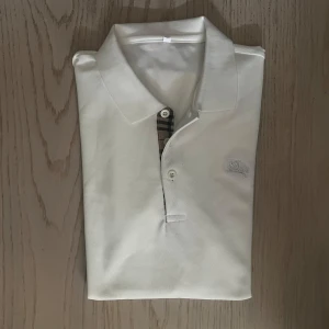 Vit Burberry t-shirt med rutig detalj - Vit t-shirt från Burberry med klassisk rutig detalj vid knäppningen och broderad logga på bröstet. T-shirten har korta ärmar, krage och är gjord i mjuk bomull. Perfekt för dig som gillar stilrena plagg med en twist.