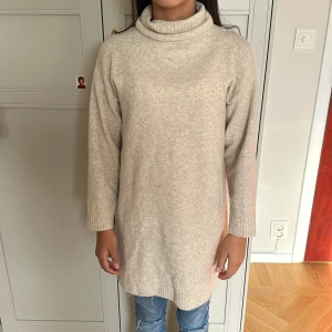 Beige stickad polotröja lång modell - Lång, beige stickad polotröja med avslappnad passform och ribbade muddar. Tröjan har hög polokrage och är perfekt att styla med jeans eller leggings. Enkel och stilren design som funkar året runt.