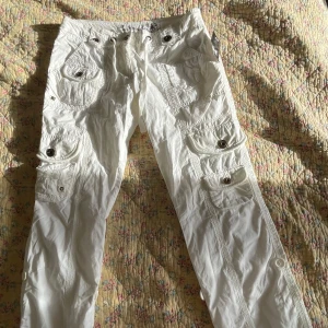 Vita cargopants med snörning - Säljer ett par vita cargopants från Sarasol med flera fickor, coola bronsfärgade knappar och snörning i midjan. Byxorna har dragkedjedetalj och är perfekta för en avslappnad streetstil.
