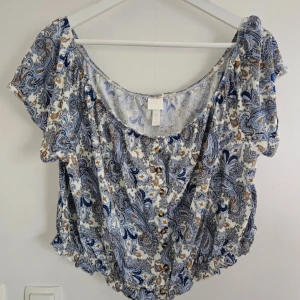 Blommig blus i blått och vitt XL - Supersöt blus med blommigt paisleymönster i blått, vitt och brunt. Modellen är croppad med korta ärmar, knappar framtill och resår i nederkant. Tunn och luftig i 100% viskos, perfekt för varma dagar. Snygg till både jeans och kjol!