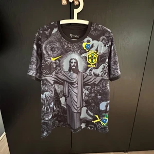 Nike Brasil svart grafisk fotbollströja - Unik svart Brasilien fotbollströja från Nike med detaljerat grafiskt tryck av Kristusstatyn och brasilianska symboler. Gula loggor och emblem på bröstet, korta ärmar och rund hals. Materialet är lätt och andas, perfekt för match eller träning.