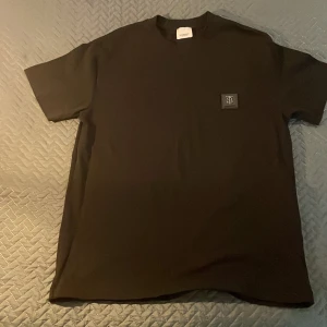 Svart Burberry t-shirt med logga - Snygg svart t-shirt från Burberry med klassisk rund hals och korta ärmar. På bröstet sitter en stilren svart patch med Burberrys TB-monogram i silver. T-shirten är i mjuk bomull och har en clean, minimalistisk look som passar till allt. Nypris 2400