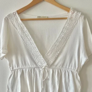 Vit omlottblus från Pull&Bear - Superfin vit blus från Pull&Bear med v-ringning och spetsdetaljer längs urringningen. Klänningen har knyt i midjan och är tillverkad i ett lätt, krinklat material. Perfekt för varma dagar och ger en fräsch och somrig vibe.