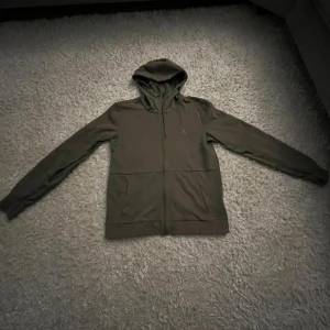 Mörkgrön hoodie med dragkedja - Säljer en stilren mörkgrön hoodie med dragkedja och huva. Tröjan har långa ärmar, två fickor framtill och ribbade muddar vid ärmslut och nederkant. Perfekt för en avslappnad look och enkel att matcha med jeans eller joggers.