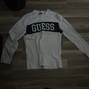 Vit långärmad tröja från Guess - Vit långärmad tröja från Guess med en bred marinblå panel över bröstet och stor vit logotyp. Tröjan har klassisk passform och rund halsringning, perfekt för en clean och sportig look.