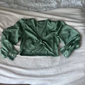 Grön kort blus med pärlknappar Zara - Snygg ljusgrön blus från Zara i glansigt satintyg. Blusen har långa puffärmar med breda manschetter och pärlknappar, samt knyt framtill. Croppad modell med v-ringning.