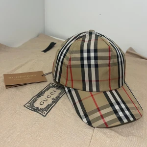 Burberry Keps - Snygg keps från Burberry med det ikoniska rutiga mönstret i beige, svart, vitt och röda linjer. Justerbar baktill med svart plastspänne och tydlig Burberry-logga. Tillverkad i bomull och polyester för en bekväm passform. Tagen gick av tyvärr och den är lite använd men exakt som ny.