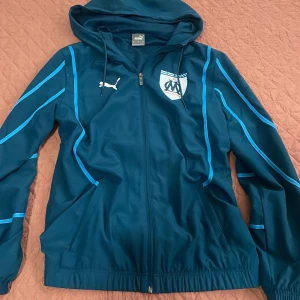 Blå vindjacka Puma Marseille S - Snygg marinblå vindjacka från Puma med Olympique Marseille-logga på bröstet. Jackan har huva med snörning, dragkedja framtill och ljusblå detaljer längs ärmarna. Perfekt för dig som gillar sportig stil och vill representera ditt lag.