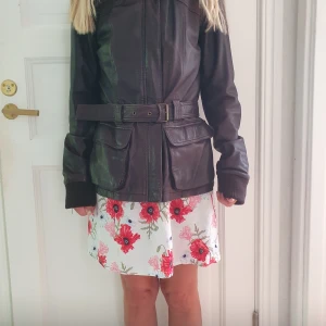 Brun skinnjacka med bälte från Zoul - Snygg mörkbrun skinnjacka från Zoul med bälte i midjan och stora fickor framtill. Jackan har långa ärmar med ribbade muddar och coola sömdetaljer. Perfekt för dig som vill ha en klassisk men edgy look.