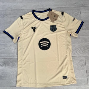  FC Barcelona borta matchtröja 25-26 - Snygg beige FC Barcelona fotbollströja från Nike med svart och blå detaljer på krage och ärmslut. Säsong 25-26