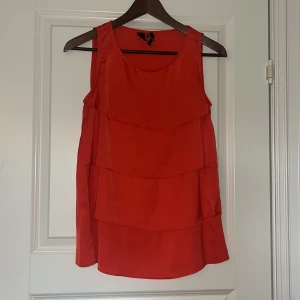 Röd volanglinne från Mango - Snygg röd ärmlös topp från Mango med flera lager volanger framtill. Toppen har rund halsringning och är tillverkad i ett glansigt, lätt material som ger en lyxig känsla. 