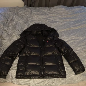 Svart pufferjacka från Polo Ralph Lauren - Säljer en svart pufferjacka från Polo Ralph Lauren med huva och dragkedja framtill. Med två sidofickor