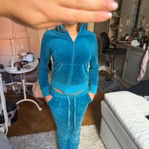 Juicy Couture blåa mjukisbyxor och hoodie - Blåa mjukisbyxor från Juicy Couture i mjuk velour med glittriga stenar längs benet. Byxorna har snörning i midjan, fickor med dragkedja och en relaxed passform. Perfekta för en chill och trendig look.