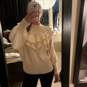 Creamvit stickad tröja med volang - Superfin stickad tröja i vit med volanger. Använd ett fåtal gånger och har inga tecken på användning. ❣️Tröjan har hög hals och ballongärmar som ger en trendig och mysig look. Perfekt för dig som gillar detaljer och vill sticka ut lite extra.💕