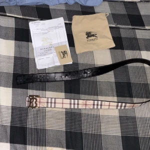 Burberry rutigt bälte med guldspänne - Säljer ett snyggt Burberry bälte med klassiskt rutigt mönster i beige, brunt och rött. Bältet har ett stort guldigt 'B'-spänne framtill och är gjort i skinn. Perfekt accessoar för att lyfta din outfit med en lyxig touch.